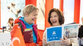 Two smiling women stand together at a stand with a Turkish flag at f.re.e and leaf through a travel magazine.