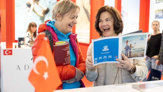 Two smiling women stand together at a stand with a Turkish flag at f.re.e and leaf through a travel magazine.