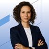Eine lächelnde Frau mit dunklen kurzen gelockten Haaren trägt ein weißes Oberteil und ein dunkles Jackett darüber.
