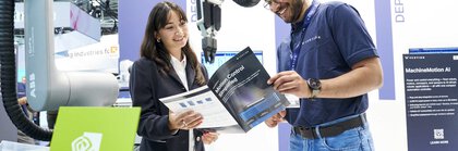 Eine Frau und ein Mann unterhalten sich lächelnd an einem Messestand über einen Prospekt mit der Aufschrift „Motion Control Simplified“. Im Hintergrund sind kollaborative Roboterarme, technische Ausstellerflächen und ein NVIDIA-Partner-Schild zu sehen.