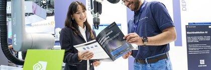A woman and a man are smiling while discussing a brochure titled “Motion Control Simplified” at a trade fair booth. In the background are collaborative robotic arms, technical displays, and a sign indicating a partnership with NVIDIA.