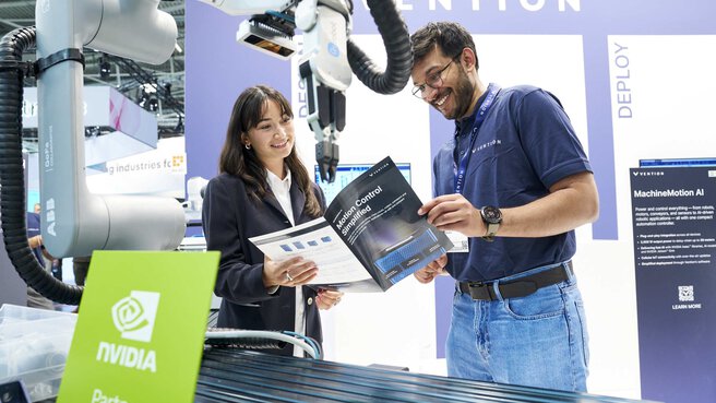A woman and a man are smiling while discussing a brochure titled “Motion Control Simplified” at a trade fair booth. In the background are collaborative robotic arms, technical displays, and a sign indicating a partnership with NVIDIA.