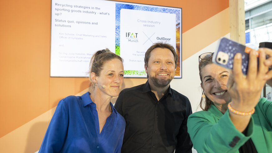 Three people take a selfie in a brightly lit room with a screen in the background.