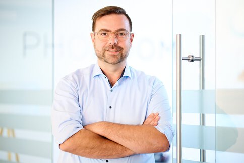 Mann mit Brille und kurzen Haaren im hellblauen kurzärmligen Hemd steht selbstbewusst mit verschränkten Armen vor einer Glastür.