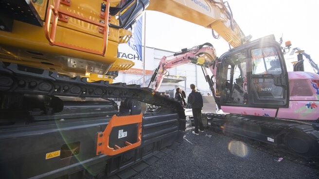 A large yellow excavator stands next to a smaller pink one; people are standing between them at a trade fair site.