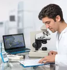 Man in lab coat looks at a sheet of paper, next to him a microscope and a laptop on which an enlargement of the sample can be seen.