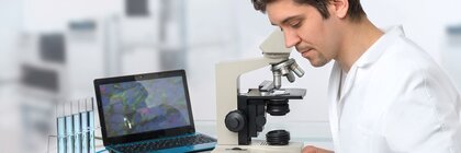 Mann im Laborkittel sieht auf ein Blatt, neben sich ein Mikroskop und einen Laptop, auf dem eine Vergrößerung der Probe zu sehen ist.