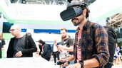 A man in a checked shirt stands at a stand with VR goggles on his head, holding two rubber handles.