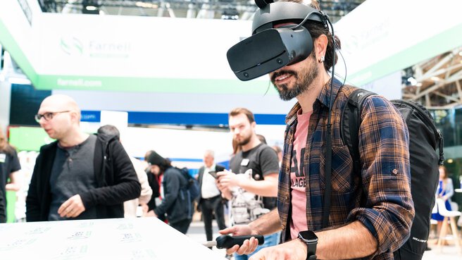 A man in a checked shirt stands at a stand with VR goggles on his head, holding two rubber handles.