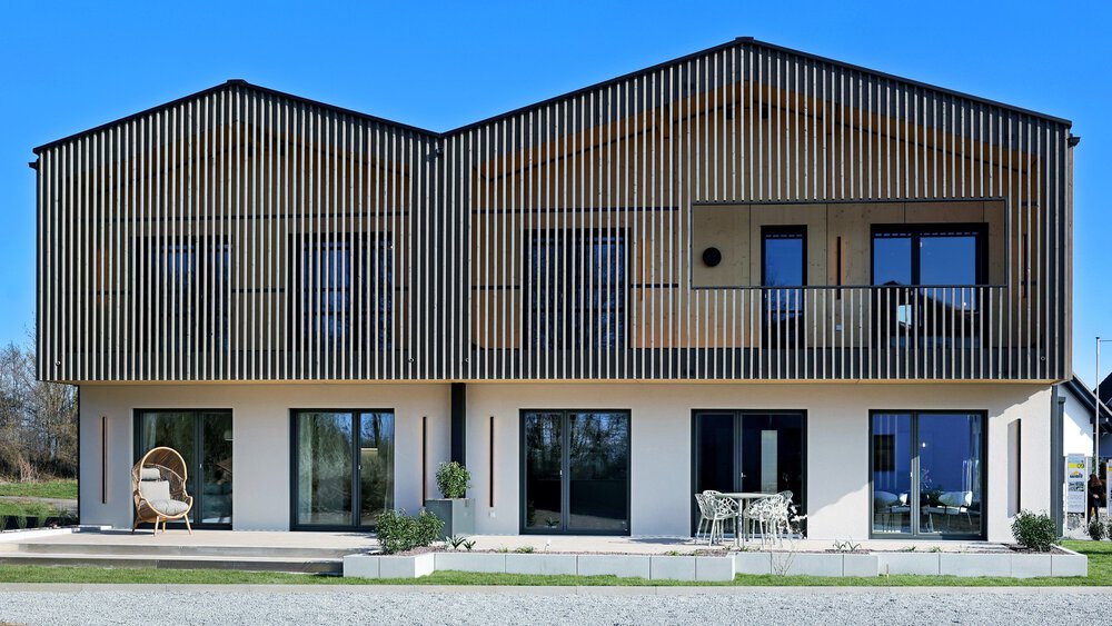 Ein modernes Haus mit senkrechten Holzlatten an der Fassade des Obergeschosses. Vor den fünf Terrassentüren befinden sich ein paar Gartenstühle und ein Tisch.