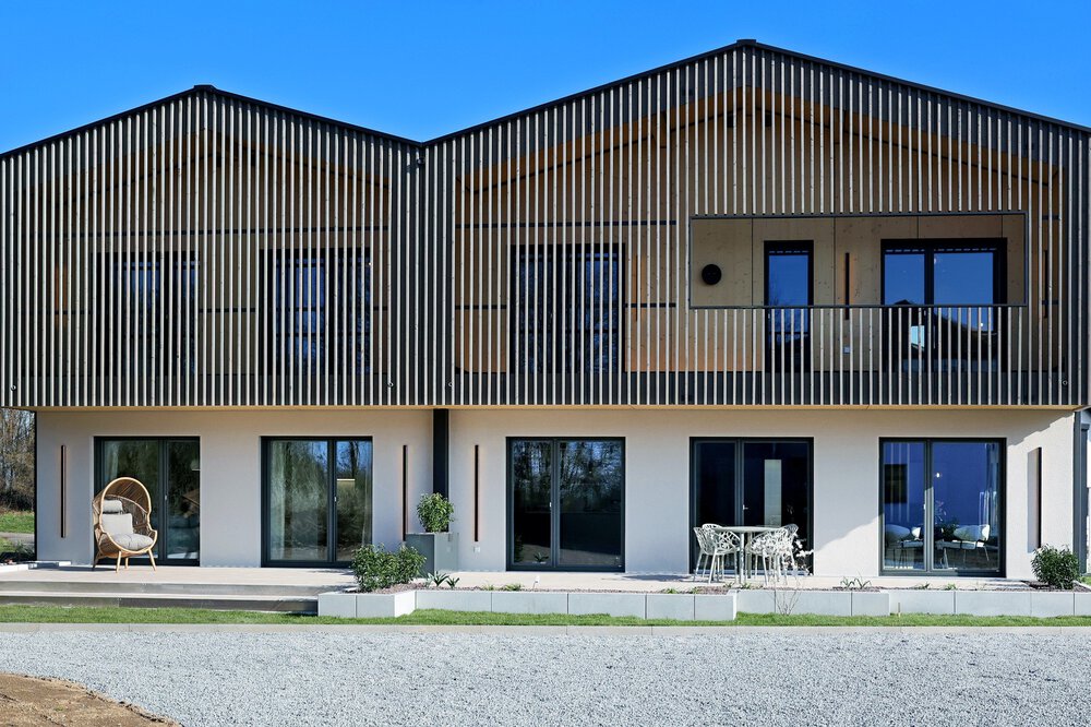 Ein modernes Haus mit senkrechten Holzlatten an der Fassade des Obergeschosses. Vor den fünf Terrassentüren befinden sich ein paar Gartenstühle und ein Tisch.