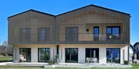 Ein modernes Haus mit senkrechten Holzlatten an der Fassade des Obergeschosses. Vor den fünf Terrassentüren befinden sich ein paar Gartenstühle und ein Tisch.