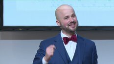 Person im Anzug und Fliege hält eine energische Rede auf einer Bühne mit Projektor-Leinwand im Hintergrund.