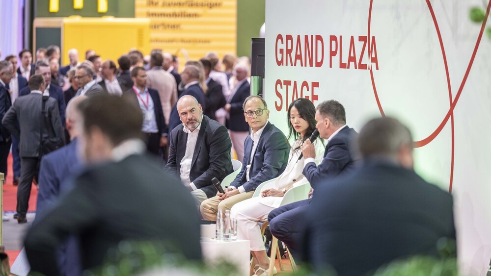 Four people with microphones are sitting on the ‘GRAND PLAZA STAGE’ at EXPO REAL, whilst numerous trade fair visitors are standing together in the background.