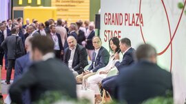 Four people with microphones are sitting on the ‘GRAND PLAZA STAGE’ at EXPO REAL, whilst numerous trade fair visitors are standing together in the background.