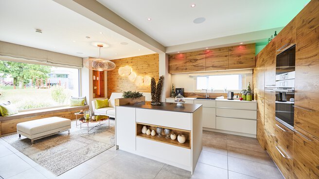 Moderne Küche und Wohnbereich mit Holzakzenten, weißen Schränken, Kochinsel und großem Fenster mit Blick auf den Garten.