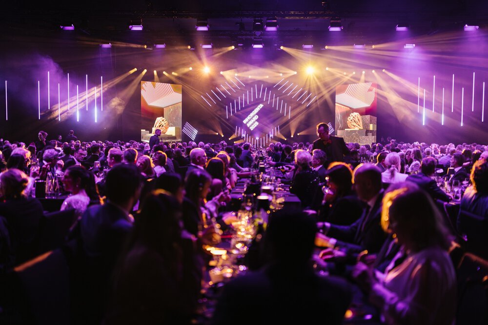 Eine große Menschenmenge bei der AWARD-Gala mit violetter Beleuchtung und Bühnenleinwänden, Leute sitzen an Tischen.