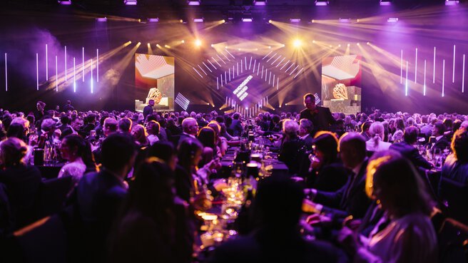 A large crowd at the AWARD Gala with purple lighting and stage screens, people sitting at tables.