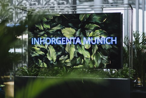 A screen with the words “INHORGENTA MUNICH” on a leaf background is surrounded by green plants.