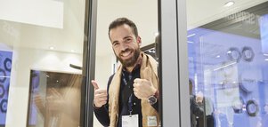 Ein Mann mit einem braunen Schal blickt aus einem geöffneten Fenster an einem Stand der BAU. Er streckt lächelnd die Daumen in die Höhe.