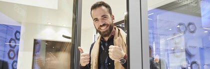 Ein Mann mit einem braunen Schal blickt aus einem geöffneten Fenster an einem Stand der BAU. Er streckt lächelnd die Daumen in die Höhe.
