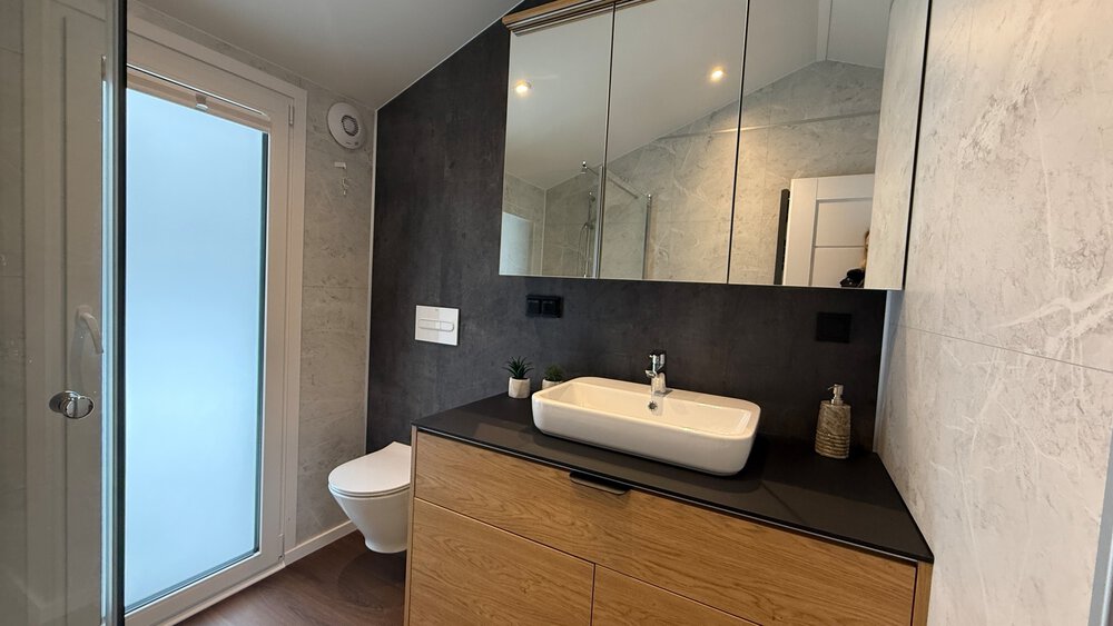 Ein Tageslichtbad mit einem weißen Waschbecken, Holzunterschrank, Spiegelschrank und einem weißen WC.