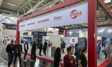 People interacting at a trade fair stand, a sign with logos of, for example, 