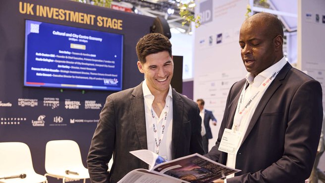 Two men in suits leaf through a brochure together. The back wall of the UK Investment Stage can be seen in the background.