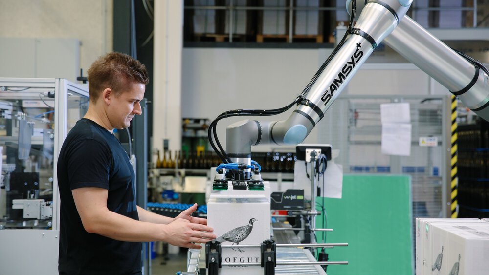 Ein Mann im schwarzen Shirt platziert eine Kiste unter einem silbernen Roboterarm, der mit einer Hebevorrichtung ausgestattet ist.