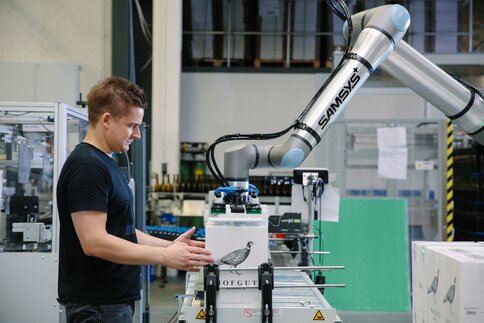 Ein Mann im schwarzen Shirt platziert eine Kiste unter einem silbernen Roboterarm, der mit einer Hebevorrichtung ausgestattet ist.