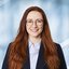 Bright smiling woman with long red hair and round glasses. She is wearing a dark blazer and a white shirt.