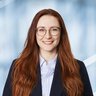 Bright smiling woman with long red hair and round glasses. She is wearing a dark blazer and a white shirt.