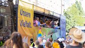 Menschen feiern auf einem Festwagen mit Regenbogenbannern, auf denen „Unsere Vielfalt“ steht, während eine Menschenmenge zusieht.