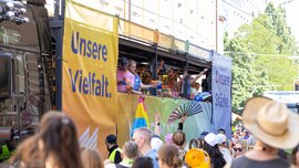 People are celebrating on a float decorated with rainbow banners that read “Our Diversity,” while a crowd looks on.