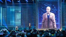 Eine Menschenmenge beobachtet einen Redner auf der Bühne, neben dem ein kleiner Roboter steht und auf einem großen digitalen Bildschirm unter blauer, futuristischer Beleuchtung ein humanoider Roboter in einem Anzug zu sehen ist.