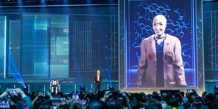 Eine Menschenmenge beobachtet einen Redner auf der Bühne, neben dem ein kleiner Roboter steht und auf einem großen digitalen Bildschirm unter blauer, futuristischer Beleuchtung ein humanoider Roboter in einem Anzug zu sehen ist.
