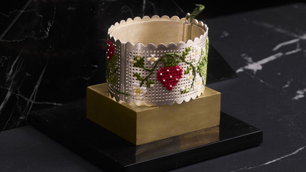 Embossed silver bracelet with a red berry and green leaf pattern, presented on a black and gold stand.