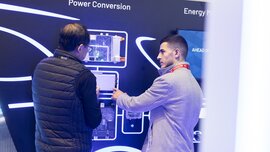 Two people are discussing in front of an exhibition wall on which various modules are mounted. The words 