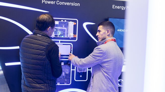 Two people are discussing in front of an exhibition wall on which various modules are mounted. The words 
