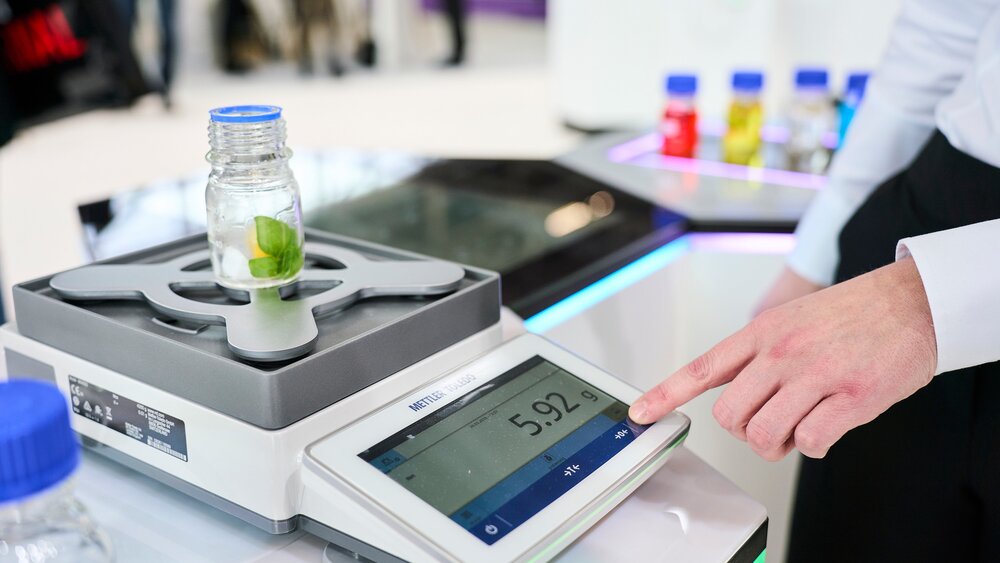 Digital laboratory scale with sample container and touchscreen controls at a trade fair stand
