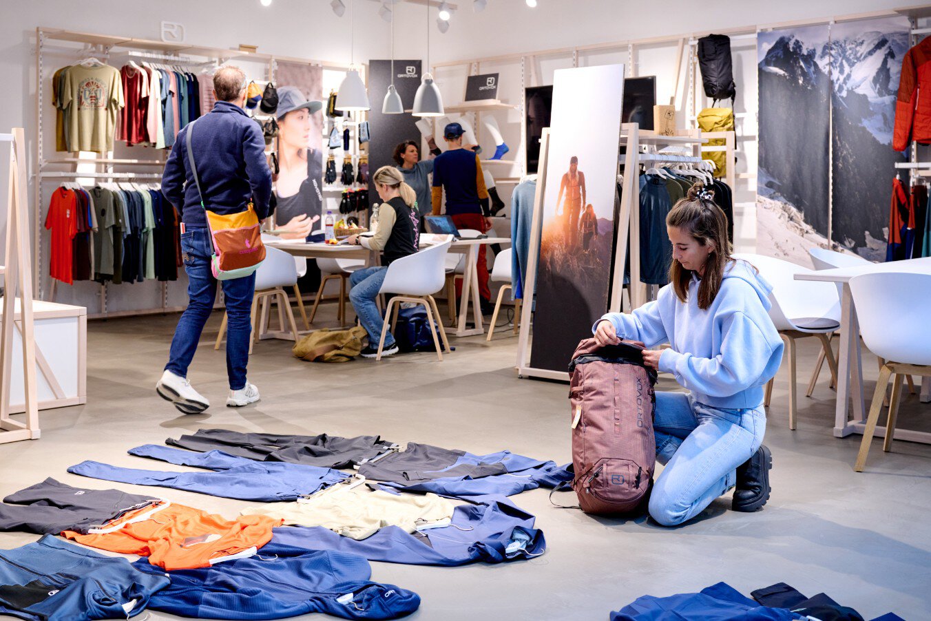 Menschen kaufen in einem modernen Geschäft Outdoor-Kleidung ein; eine Frau inspiziert einen Rucksack auf dem Boden.