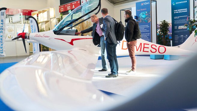 A small white and red aeroplane stands on a stand in an exhibition hall at productronica. Three men are looking at the inside of the machine.