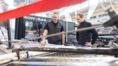 Zwei Personen befinden sich an einem Messestand für Wassersport und begutachten gemeinsam ein ausgestelltes Segelboot. Im Hintergrund sind weitere Messestände und Werbeflächen der Aussteller zu sehen.