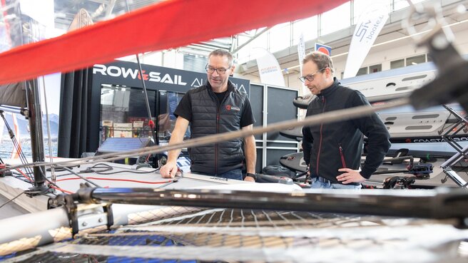 Zwei Personen befinden sich an einem Messestand für Wassersport und begutachten gemeinsam ein ausgestelltes Segelboot. Im Hintergrund sind weitere Messestände und Werbeflächen der Aussteller zu sehen.