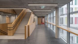 Ein großer leerer Flur mit Betonstützen und -decke grenzt an ein holzvertäfeltes Treppenhaus. In der rechten Wand sind Fensterfronten eingelassen.
