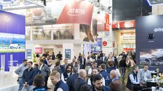 Numerous visitors to transport logistic stand between several exhibition stands and chat. Advertising banners from various exhibitors hang above their heads.