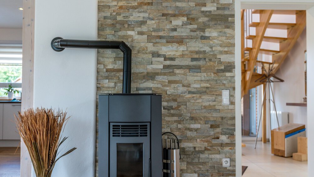 Ein moderner Holzofen vor einer Steinwand, neben einer Treppe und einigen getrockneten Pflanzen in einer Vase.