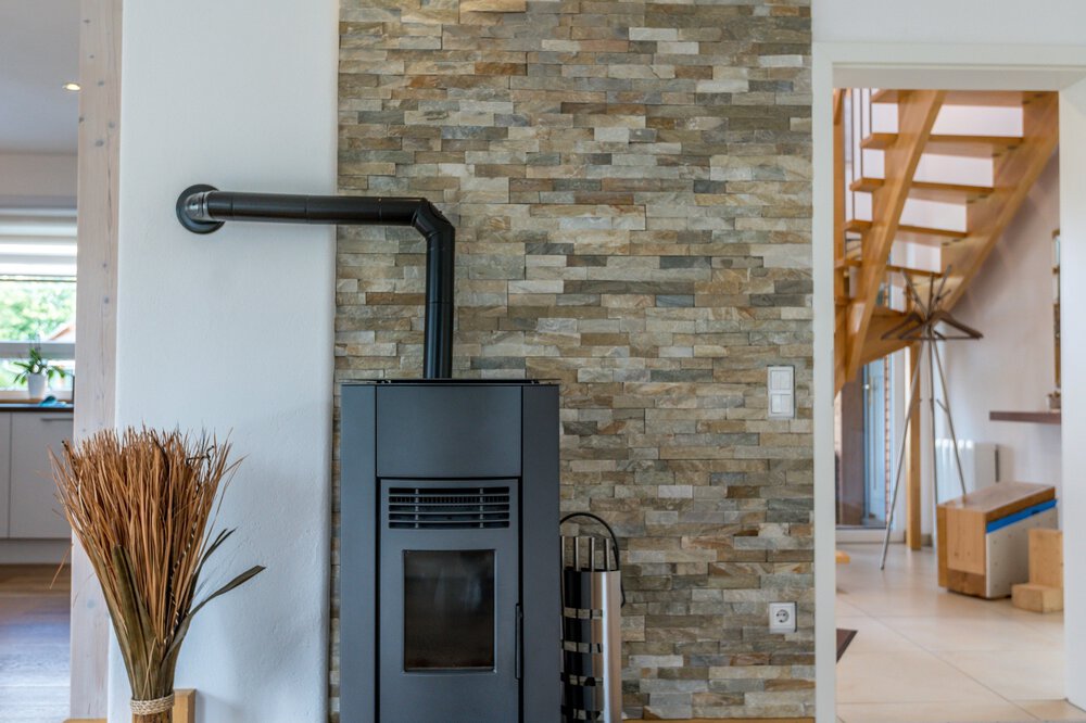 Ein moderner Holzofen vor einer Steinwand, neben einer Treppe und einigen getrockneten Pflanzen in einer Vase.