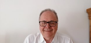 Ein Mann mit schwarzer Brille und weißem Hemd steht lächelnd vor einer weißen Wand.
