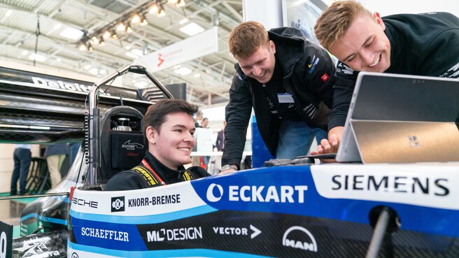 A man is sitting in a racing car that is on display at a trade fair stand. Two other men are leaning over the vehicle and typing on a tablet.